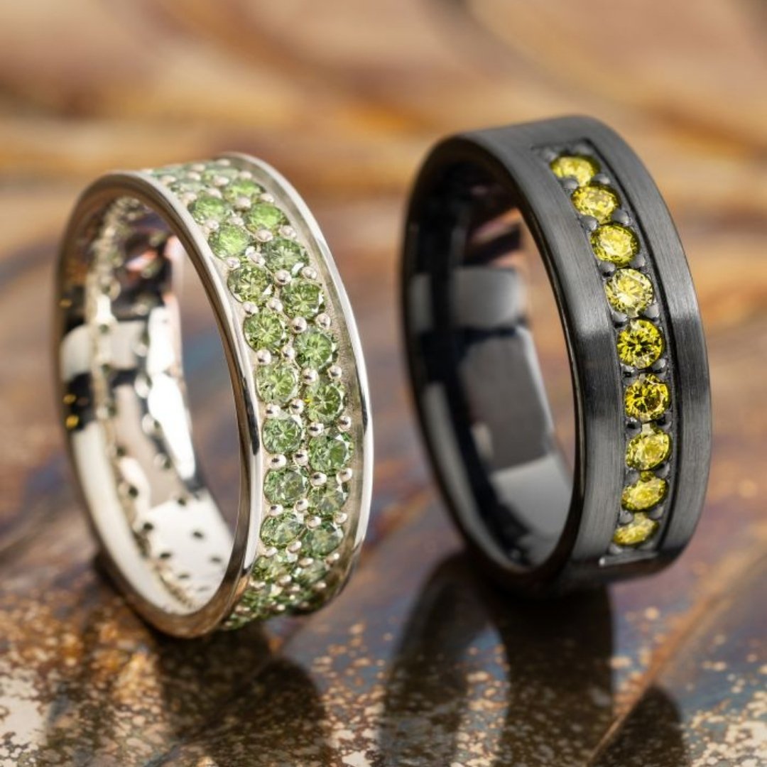 Two make wedding bands, one white gold with green stones and with a dark metal and yellow stones.