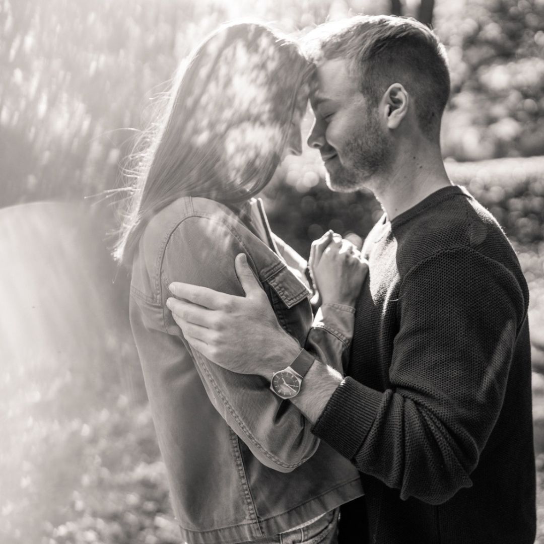 Black and white photo of a couple embracing outdoors with sunlight filtering through after getting engaged.
